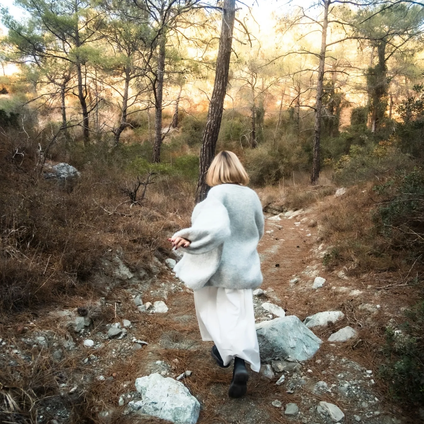 A candid moment of a woman moving freely through a woodland path, wearing light, flowing clothes that match the soft golden tones of the forest. The image evokes a sense of freedom, mindfulness, and connection with nature.